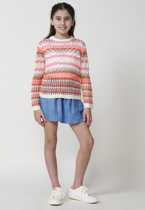Pull en tricot avec un motif à rayures multicolores dans les tons de rose, orange et blanc, associé à un short en denim bleu et des baskets blanches.