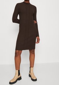 Femme portant une robe marron côtelée à manches longues et longueur genou, associée à des bottines à semelle épaisse beige, debout devant un fond uni.