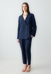 Granatowy lniany żakiet podwójnie zapięty z dopasowanymi spodniami zwężanymi, z wykładanymi klapami i dwiema przednimi kieszeniami; w komplecie z beżowymi sandałami na obcasie.