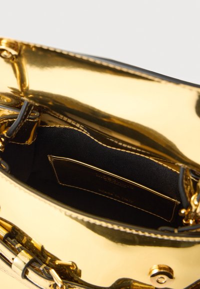 Ανοιχτή γυαλιστερή χρυσή τσάντα χειρός που αποκαλύπτει εσωτερική ύφανση από μαύρο ύφασμα με δερμάτινη ετικέτα ραμμένη στο εσωτερικό.