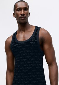 Top negru fără mâneci, cu un model texturat de logo-uri. Materialul pare neted, având un design cu spate tip racer și o linie de gât contrastantă.