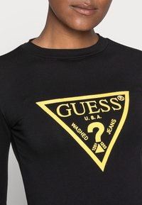 Schwarzes Langarmshirt mit einem gelben dreieckigen Logo, auf dem der Text "GUESS U.S.A. WASHED JEANS" und ein Fragezeichen in der Mitte zu sehen ist.