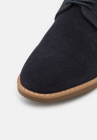 Chaussure en daim bleu foncé avec un bout arrondi, semelle en caoutchouc beige, et coutures contrastantes le long du bord. Langue texturée et lacets.