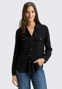 Black long-sleeve shirt with button-up front, two chest pockets, and silver button accents. Smooth fabric, collared neckline, and relaxed fit.