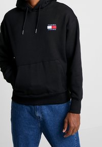 Felpa nera in cotone, con tasca frontale a sacca, cappuccio regolabile con corde e logo ricamato "TOMMY JEANS" sul petto.