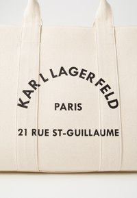 Beredskapstygsax i beige med robusta handtag. Har djärv svart text: "KARL LAGERFELD PARIS 21 RUE ST-GUILLAUME" i en böjd arrangemang.