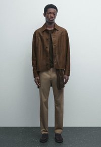 Brązowa skórzana kurtka na ciemnej bluzce, w połączeniu z beżowymi spodniami i ciemnymi butami. Kurtka ma zapięcie na guziki i luźny krój.