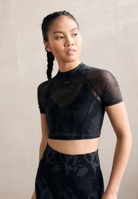 Top deportivo negro cortado con mangas cortas, que presenta un diseño de malla transparente y un patrón sutil, combinado con leggings negros de cintura alta.