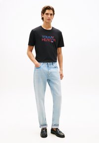 Μαύρο μπλουζάκι με λογότυπο "TOMMY HILFIGER" σε κόκκινο και μπλε, συνδυασμένο με ανοιχτό μπλε τζιν κανονικής γραμμής και μαύρα loafers. Ομαλή υφή βαμβακιού.