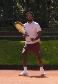 Tennisimängija heledas roosa polos ja pruunides lühikestes pükstes, hoides kollaste triipudega reketti punasel savi väljakul rohelise muru taustal.