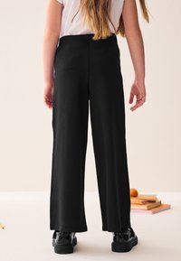 Musta, leveälahkeiset housut pehmeästä kankaasta, jossa on sileä pinta ja pehmeä laskeutuvuus. Yhdistetty kiiltävän mustien kenkien kanssa.