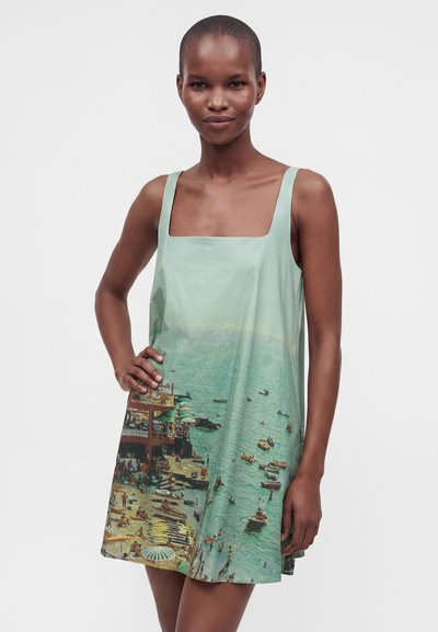 Femme mince portant une robe sans manches avec une scène imprimée de plage côtière comprenant de l'eau, des bateaux, des parasols et des personnes sur un fond clair.