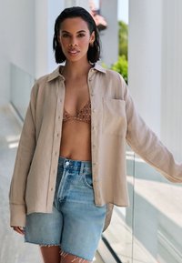 Light beige linen shirt, buttoned, with a chest pocket, over a patterned brown bikini top. Blue denim shorts with frayed hem.