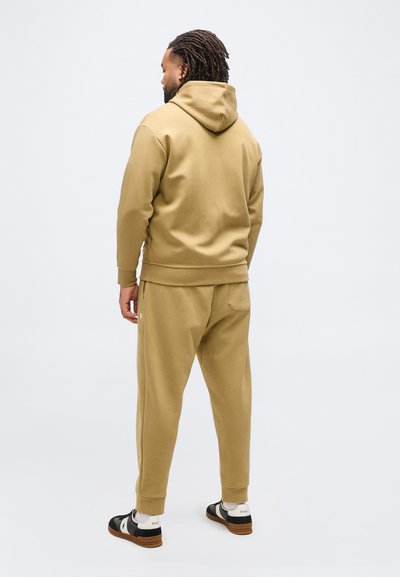 Άνδρας που στέκεται πλάτη, φοράει μπεζ φούτερ με κουκούλα, ταιριαστά παντελόνια φόρμας, λευκές κάλτσες και μαύρα-λευκά αθλητικά παπούτσια με καφέ σόλες.