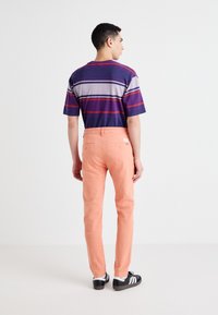 Striped short-sleeve shirt in navy, purple, and red; paired with fitted peach trousers; black shoes; viewed from the back.