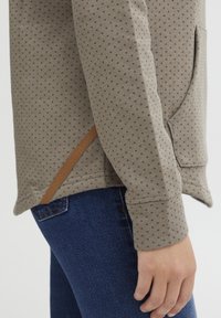 Pullover color marrone chiaro con un motivo a pois, dotato di una tasca laterale e di un'etichetta del marchio marrone sull'orlo; abbinato a jeans blu scuro.