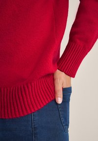 Suéter de punto rojo con puños y dobladillo acanalados, que presenta una textura suave. Combinado con unos vaqueros de mezclilla azul oscuro. Primer plano de una mano en el bolsillo.