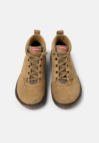 Zapatos de ante marrón con acentos grises, punta redonda y suela de goma marrón. Presentan un sistema de cordones en color beige y un logo rojo de "Camper" en la lengüeta.