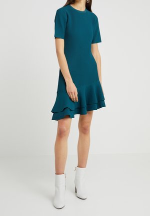 Kobieta w zielonkawoniebieskiej, krótko rękawowej sukience prążkowanej z asymetrycznym, warstwowym falbanowym dołem oraz białych botkach, stojąca na jednolitym tle.