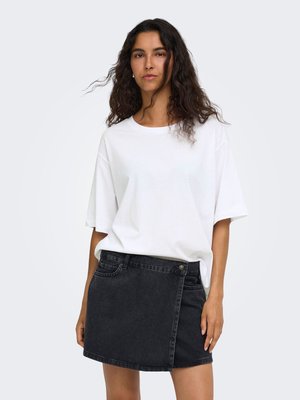 Femme aux longs cheveux bouclés et foncés, portant une chemise blanche ample à manches courtes et une jupe portefeuille en jean noir, debout devant un fond clair uni.