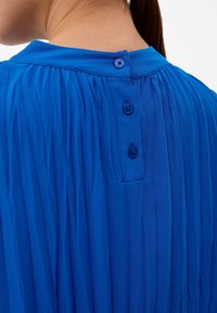 Top plissettato blu con colletto arrotondato e tre distinti bottoni sul retro. Tessuto liscio con colore vivace.