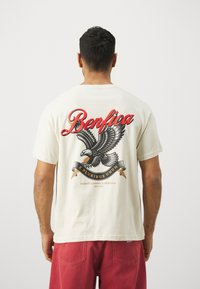 Krēma krāsas kokvilnas T-krekls ar lielu ērgļa grafiku un sarkano tekstu "Benfica", kā arī dekoratīviem baneriem.