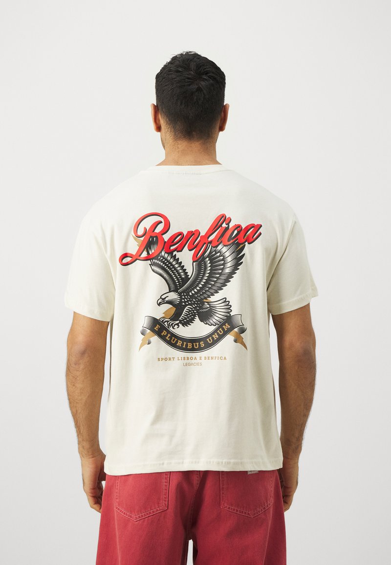Krēma krāsas kokvilnas T-krekls ar lielu ērgļa grafiku un sarkano tekstu "Benfica", kā arī dekoratīviem baneriem.