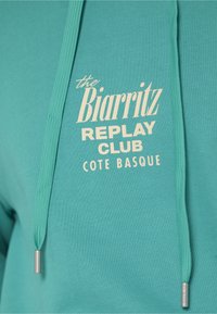 Sweat à capuche turquoise avec cordons affichant un texte crème : « the Biarritz REPLAY CLUB COTE BASQUE » sur la poitrine.
