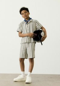 Set di maglietta a maniche corte e pantaloni corti a righe in beige e blu navy, in misto cotone, con maniche arrotolate; tiene un casco nero in una mano.