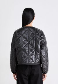 Vrouw met lang zwart haar in een gewatteerde zwarte jas met geometrische en cirkelvormige stiksels, gezien vanuit de achterkant tegen een effen achtergrond.