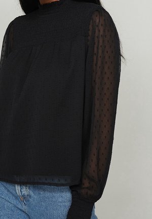 Schwarze, strukturierte Bluse mit hohem Kragen, transparenten langen Ärmeln und elastischen Bündchen, getragen zu blauen Denim-Jeans vor neutralem Hintergrund.