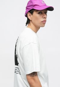 Λευκό T-shirt με γραφικό τύπωμα στη πλάτη και μαύρο κείμενο. Ανοιχτό μοβ καπέλο με λογότυπο στο μπροστινό μέρος. Κανονική εφαρμογή και εμφανής υφή.