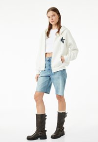 Joven mujer vestida con un cropped hoodie blanco, shorts de mezclilla azul claros y botas marrón oscuro hasta la mitad de la pantorrilla con hebillas, de pie contra un fondo blanco.