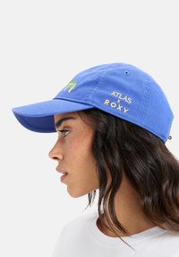 Blue cotton cap features a curved visor, embroidered 'Atlas x Roxy' logo, and a green and yellow graphic design. Soft, textured fabric.