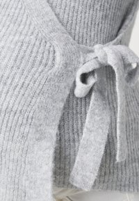 Pull en tricot gris avec une texture côtelée, un design à l'avant ouvert et un détail en nœud à gauche. Matière douce et confortable.
