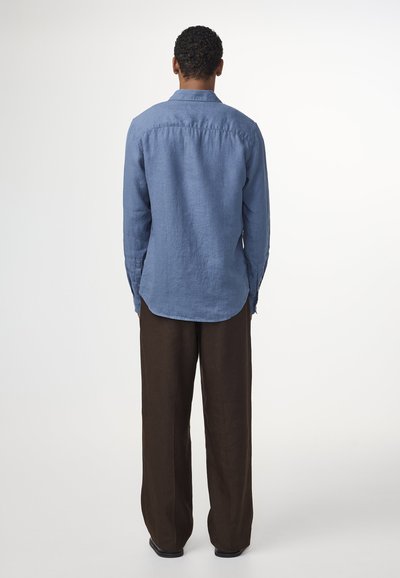 Light blue long-sleeve shirt in linen with a curved hem, paired with loose-fitting dark brown trousers, viewed from the back.