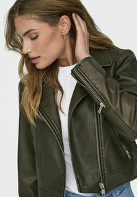Giacca da moto in pelle verde oliva con zip argentate, reverse strutturate e design aderente. Indossata sopra un top bianco, con dettagli di cucitura visibili.