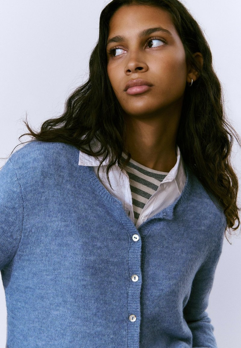 Cardigan azul claro hecho de una tela suave, con cuello en V, cinco botones y puños acanalados. Lleva puesto sobre una camisa a rayas.