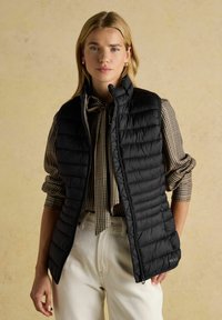 Fekete steppelt pufi mellény magas gallérral, kockás inggel és krémszínű nadrággal párosítva, cipzárral és oldalsó zsebekkel.