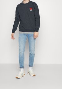 Sudadera gris oscuro con un logo bordado de la letra 'W' en rojo y verde, combinada con jeans ajustados de color azul claro y zapatillas crema.