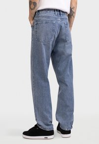 Ανοιχτό μπλε τζιν με άνετη γραμμή, διαθέτει δύο πίσω τσέπες και ραμμένες λεπτομέρειες. Συνδυασμένο με μαύρα αθλητικά παπούτσια.