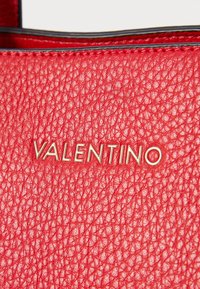 Raudona odinė rankinė, turinti tekstūruotą paviršių ir auksinę "VALENTINO" logotipą. Papuošta juodais siūlais palei kraštus.