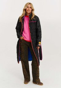 Čierny prešívaný kabát s jasne farebným lemovaním, oblečený na ružovom svetre a hnedých nohaviciach s leopardím vzorom. Hnedé kĺzavé topánky.