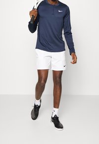 Tummansininen pitkän hihan urheilupaita, jossa on puolivälin napitus, yhdistetty valkoisiin shortseihin, joissa on taskut ja pieni musta Nike-logo. Mustat kengät.