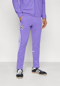 Μωβ αθλητικό παντελόνι από απαλό ύφασμα, με λευκές ρίγες στα πλάγια και λογότυπο της Ρεάλ Μαδρίτης στο πάνω μέρος του ποδιού. Συνδυασμένο με μαύρα αθλητικά παπούτσια.