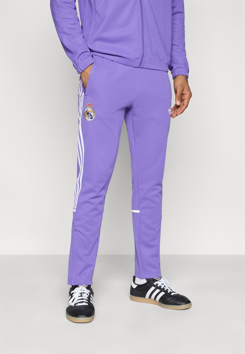 Μωβ αθλητικό παντελόνι από απαλό ύφασμα, με λευκές ρίγες στα πλάγια και λογότυπο της Ρεάλ Μαδρίτης στο πάνω μέρος του ποδιού. Συνδυασμένο με μαύρα αθλητικά παπούτσια.