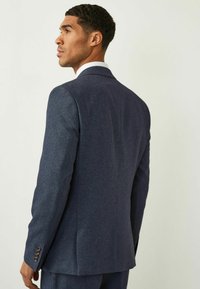 Veste de costume bleu marine texturée avec une coupe ajustée, fente unique et accents de boutons à quatre boutons. La vue arrière montre des lignes épurées et un motif subtil.