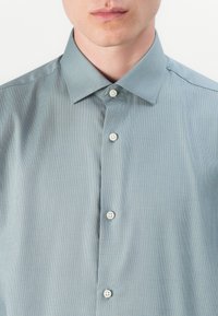 Camisa abotonada de un tono verde claro con un fino patrón a cuadros. Cuenta con un cuello clásico y cinco botones blancos en el frente. Textura suave.
