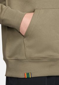 Sweatshirt verde azeitona com punhos canelados, bolso lateral e uma etiqueta colorida listrada na bainha. Material macio e corte solto.