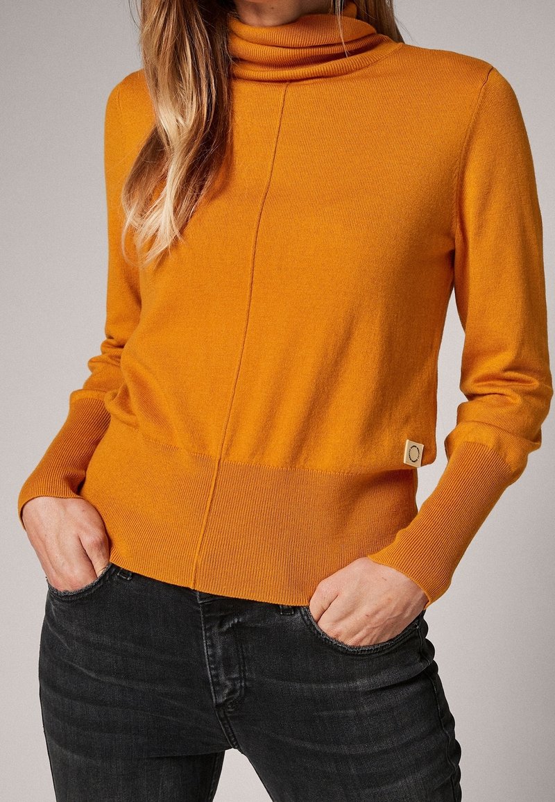 Maglione arancione con collo a cappuccio, texture a coste e design aderente. Presenta un'orlo basso e un piccolo logo ricamato. Indossato con jeans scuri.